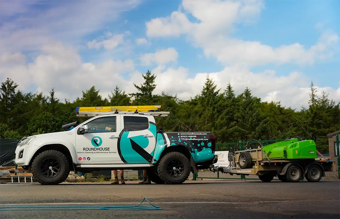Roundhouse UK branded pickup truck towing a green exterior cleaning machine on a trailer, parked at a job site with trees in the background.