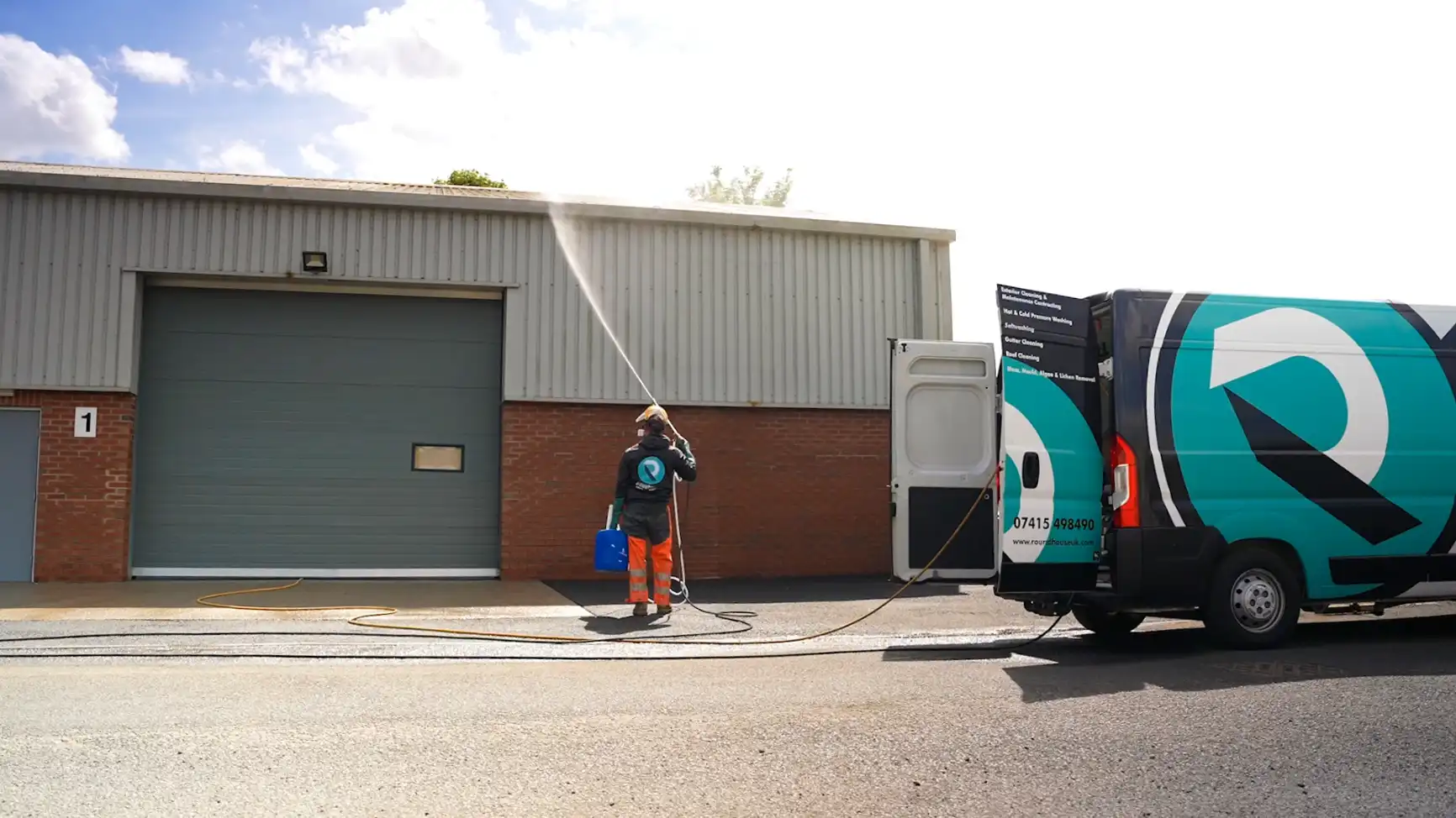 Roundhouse UK technician using an 8 m telescopic lance to soft-wash grey steel cladding on an industrial unit, service van parked beside him.