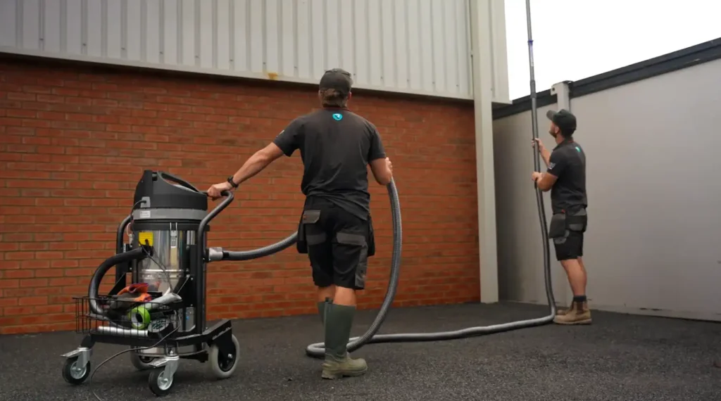 Two Roundhouse UK technicians operate a wheeled SkyVac industrial vacuum while one vacuums a high warehouse gutter with a telescopic carbon-fibre pole.