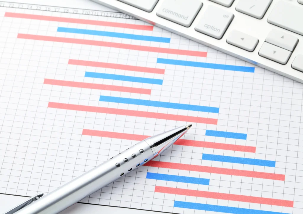 Close-up of a Gantt chart with a silver pen and computer keyboard, representing technology-driven planned preventative maintenance scheduling.