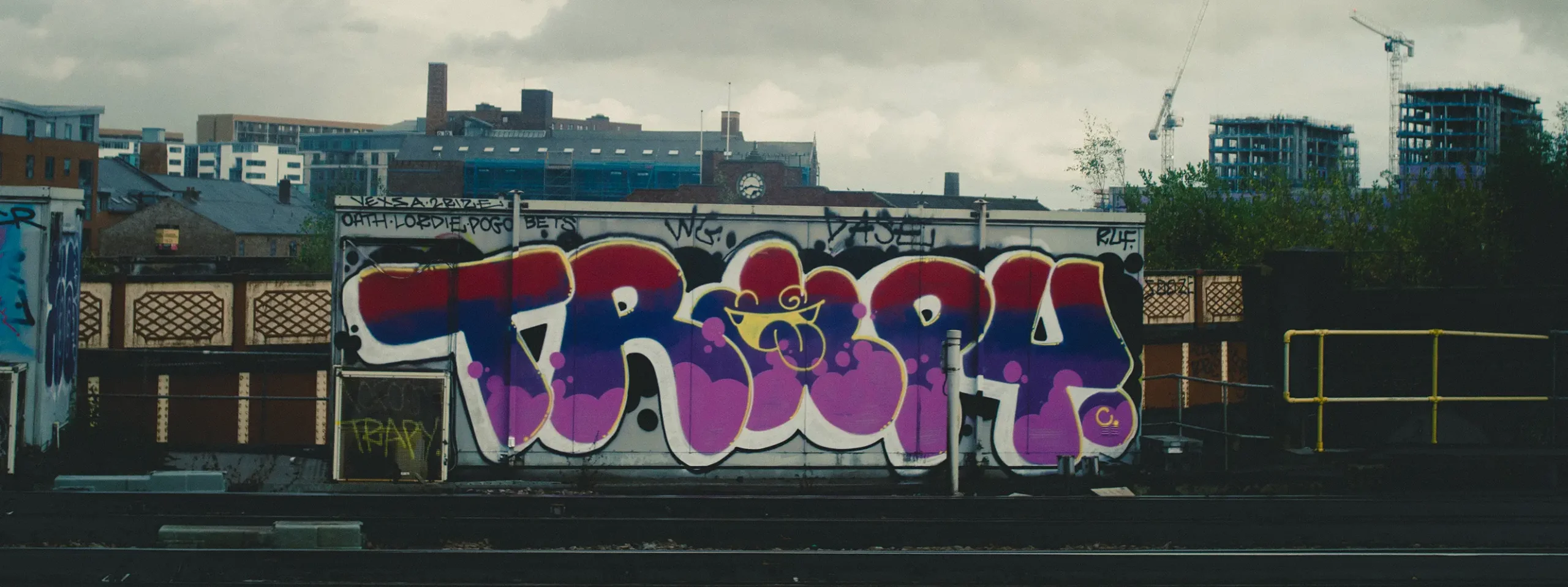 Graffiti covering a railway wall with city buildings and cranes in the background.